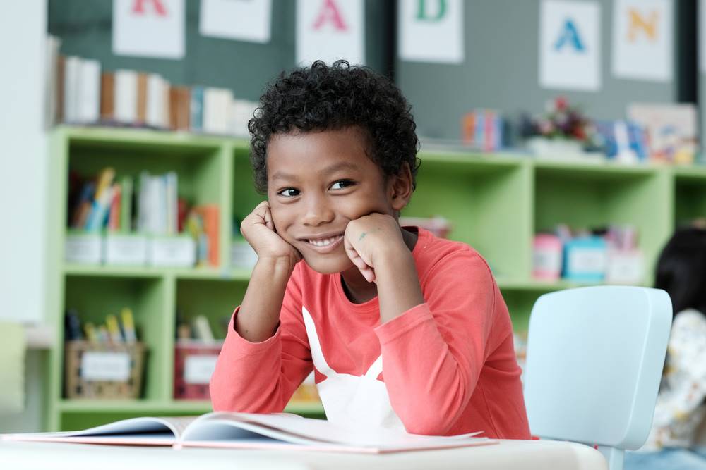 A young boy with curly hair, wearing an orange shirt, sitting at a desk with an open book in front of him. He is smiling and resting his chin on his hands, looking content. The background shows a classroom with bookshelves and educational materials.
