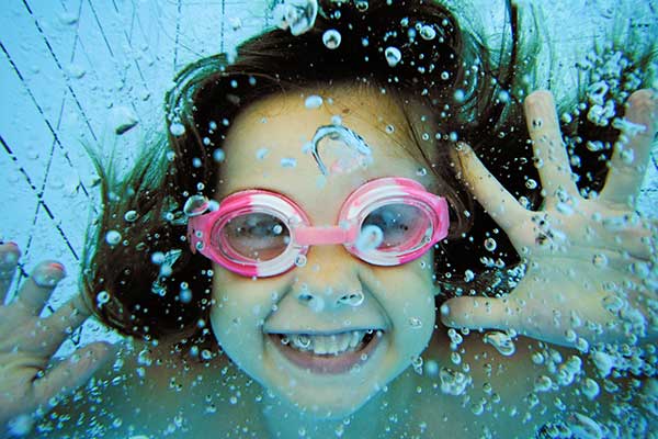 girl wearing swim goggles underwater waving at the camera