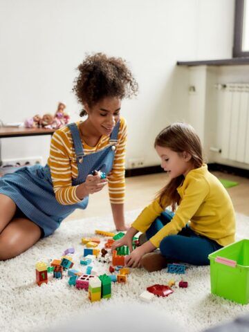 A young woman sits on the floor with a little girl who is playing with blocks