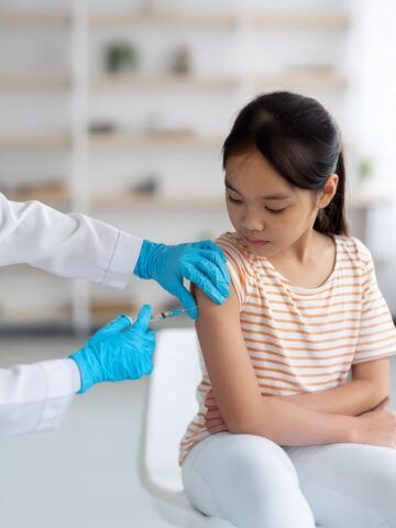 Doctor hands in protective gloves making vaccination against coronavirus of flu for little asian girl, home interior, closeup shot. Kids immunization against various disease concept
