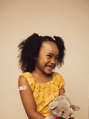 Cute girl with bandage on arm after getting a vaccine smiling. Girl holding her teddy bear looking away and smiling after vaccination.