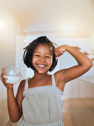 Milk, portrait and African girl with muscle from healthy drink for energy, growth and nutrition in the kitchen. Happy, smile and child flexing muscles from calcium in a glass and care for health