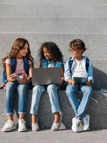 Smiling school kids preteen boys and girls elementary middle pupils children sitting on the stairs and looking in smartphone screen with digital devices laptop tablet in hands outdoors, copy space